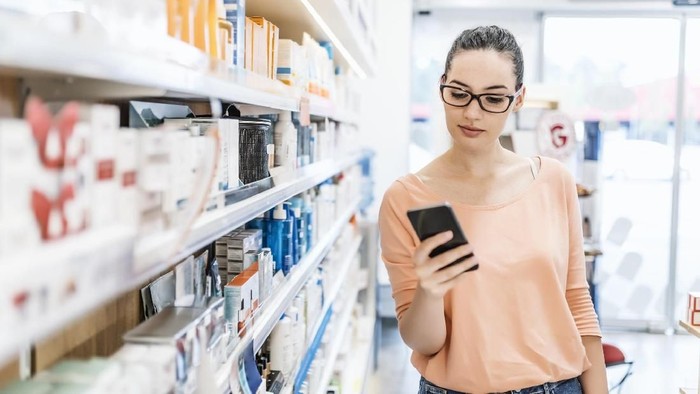 Photo of female Caucasian young consumer checking information on mobile phone in pharmacy. Medicine, pharmaceutics, health care and people concept. Woman using black smartphone while shopping in the pharmacy store during the day.