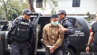 Tampak Hermawan mengenakan jaket warna cokelat, peci hitam, dan celana panjang warna abu-abu. Hermawan juga mengenakan masker warna hijau.