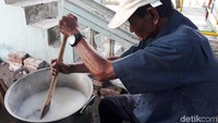Seorang warga tengah duduk di depan panci berukuran besar berisi beras dan air. Sembari mendengarkan alunan lagu era jaman dahulu, pria bertopi ini dengan santai mengaduk rendaman beras dan air di panci tersebut.