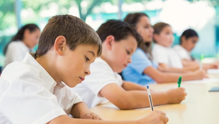 Elementary age Hispanic little boy is sitting at desk with classmates in private elementary school. He is using a pen to write on paper during exam. Classmates are also concentrating while taking test. Students are wearing white and blue school uniforms.