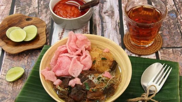 Soto Padang, the traditional beef jerky soup from Padang, West Sumatra. Crispy beef jerky with potato patty and glass noodles served in clear, spicy beef broth. Accompanied with pink tapioca crackers. The soup is plated in a ceramic bowl and placed on a plate lined with banana leaf. A set of rustic cutlery is placed on the plate.