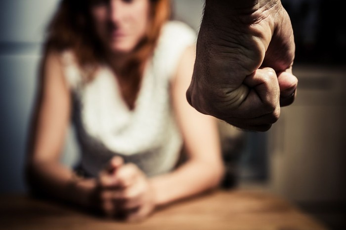 ilustrasi kekerasan dalam rumah tangga Young woman is sitting hunched at a table at home, the focus is on a mans fist in the foregound of the image