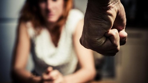 Young woman is sitting hunched at a table at home, the focus is on a mans fist in the foregound of the image