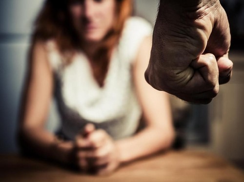 Young woman is sitting hunched at a table at home, the focus is on a mans fist in the foregound of the image