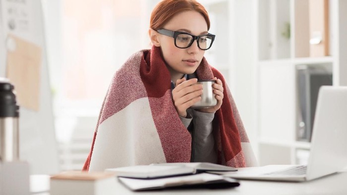 Young woman in plaid and eyeglasses drinking hot tea from mug during work in office