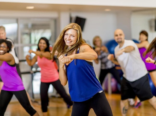A woman is leading a multi-ethnic boxing class, everyone is wearing athletic clothing and is throwing a punch together - they are smiling and looking at the camera.