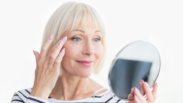 Happy senior woman applying eye anti-wrinkle cream, looking at mirror
