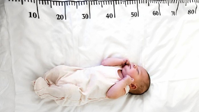 A three month baby in mint green clothes lying on a bed on which a measuring ruler for growth is drawn. Teething tool in hands, chewing