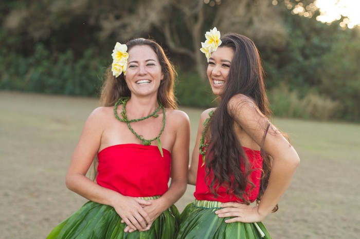 Two beautiful young local women practice hula on a beautiful day outside. Theyre standing together and laughing together at something happening off-camera.