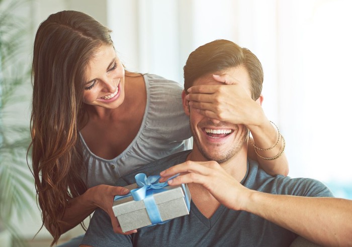 Young female keeps eyes closed to his boyfriend at home, while he is sitting on sofa with gift box, celebrating birthday