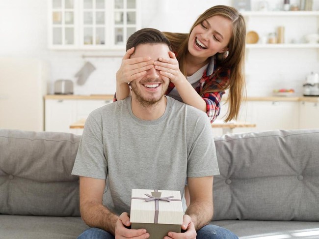 couple Young female keeps eyes closed to his boyfriend at home, while he is sitting on sofa with gift box, celebrating birthday