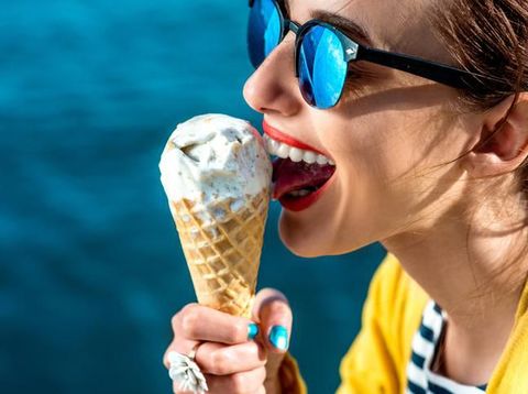 Young woman in yellow sweater and sunglasses eating ice cream on the blue water background