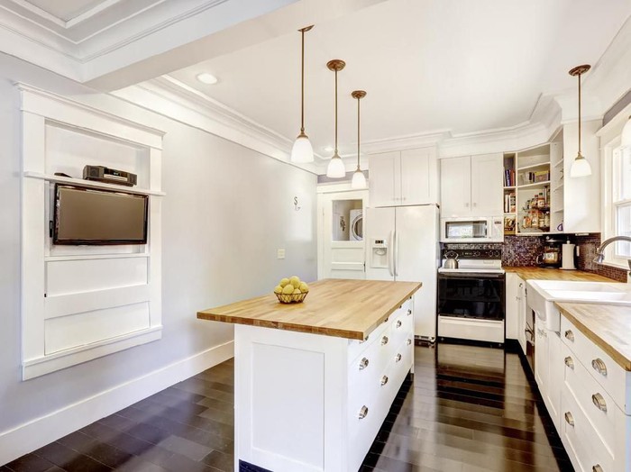 Kitchen interior in white tones with hardwood counter tops, kitchen island and black tile back splash trim. White coffered ceiling with pendant lights make room more elegant.