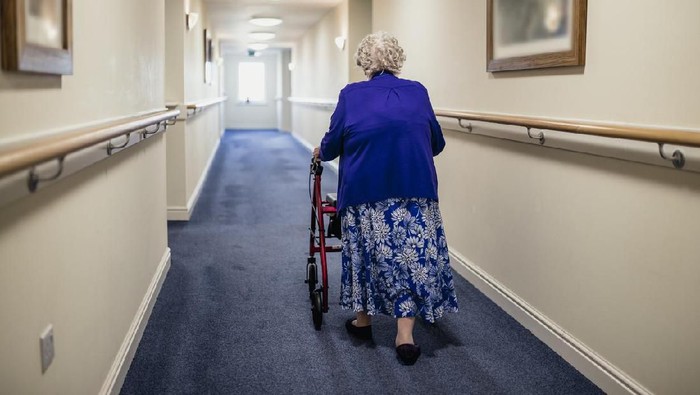 A senior woman walking down a corridor with the assistance of a walker. view from rear