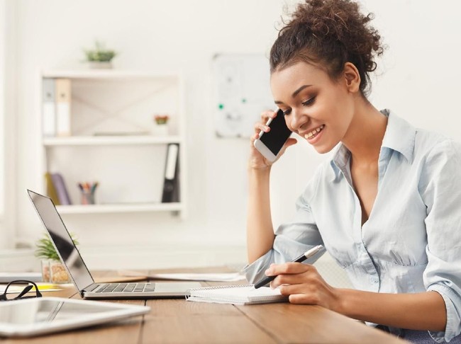 Smiling african-american business woman at work talking on phone and taking notes, sitting at her working place in office, copy space
