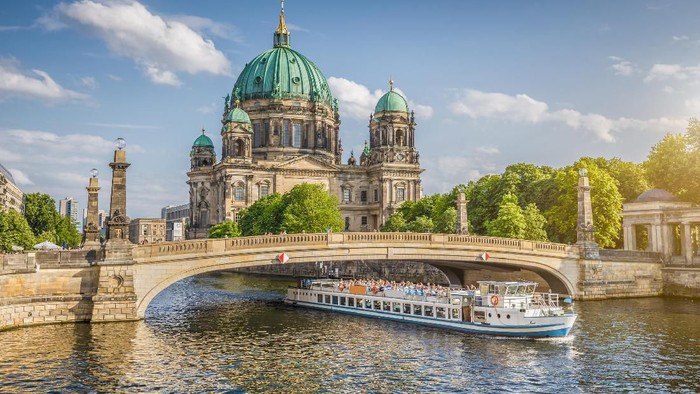 Beautiful view of historic Berlin Cathedral (Berliner Dom) at famous Museum Island with ship passing Friedrichsbrucke bridge on Spree river in golden evening light at sunset in summer, Berlin, Germany