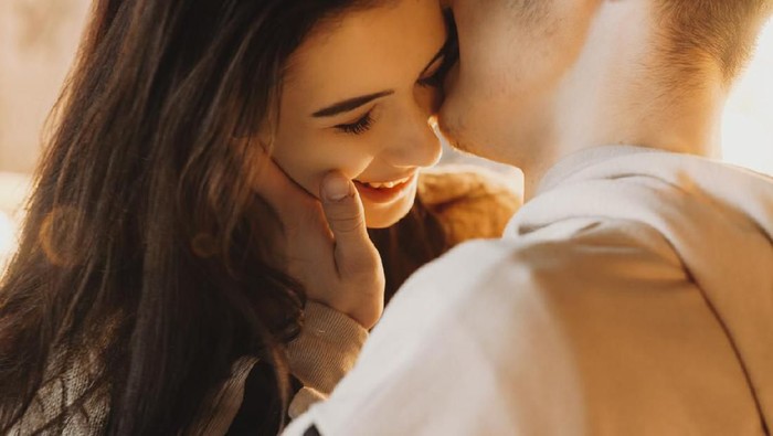 Beautiful close up portrait of an caucasian young couple embracing against light, while boy is kissing his girlfriend cheek which is smiling.