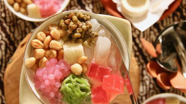 Wedang Angsle, the traditional Javanese warm dessert from Malang, East Java. Tapioca pearls, rice noodle cake, agar jelly cubes, palm fruit, mung beans, peanuts and bread cubes in warm infusion of ginger and coconut milk. The dish is plated in a ceramic deep bowl with a metal soup spoon inserted in the bowl. A batik cloth is used as a table cloth.