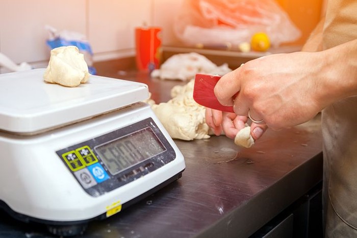 resep masakan A close-up of a female baker weighs on kitchen scales the dough for cooking buns on a knotted table with a rolling pin
