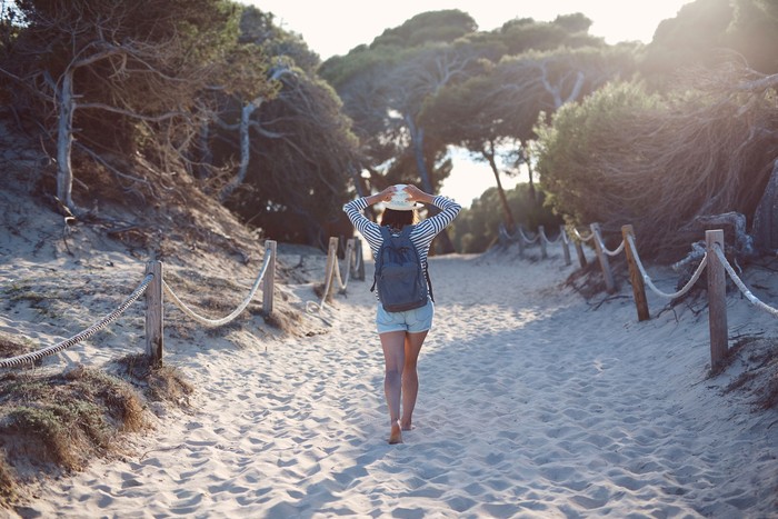 A photo of young woman walking on sandy footpath amidst plants in the summer. She is wearing backpack, holding her hat with a palm, backlit by setting sun.
