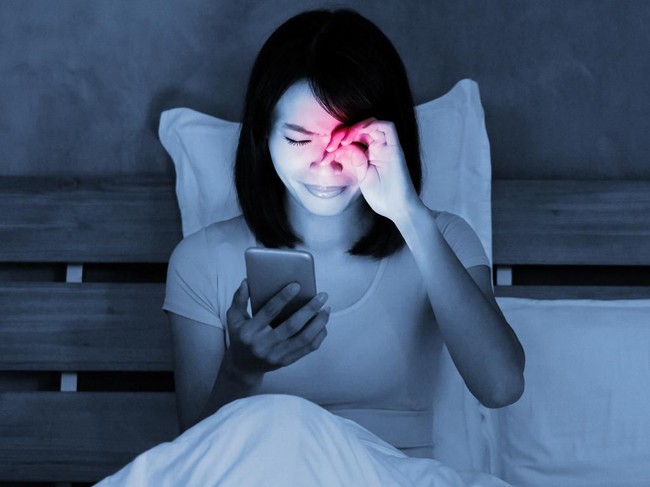 Tired woman with headache in bed with smartphone, low light
