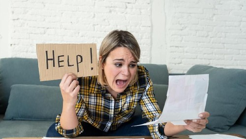 Lifestyle portrait of worried desperate young woman feeling stressed while working through finances at home showing Help sign. In single mother paying off debts and bills and financial problems.