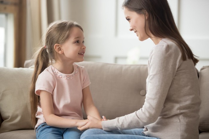 ucapan ortu Loving family happy mother and cute child girl holding hands talking sitting on sofa at home, caring elder sister mom baby sitter having friendly trust conversation with preschool little kid daughter