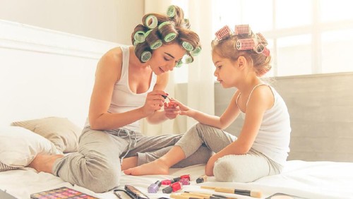 Beautiful young mother and her daughter with hair curlers are sitting on bed at home. Mom is doing her daughter manicure