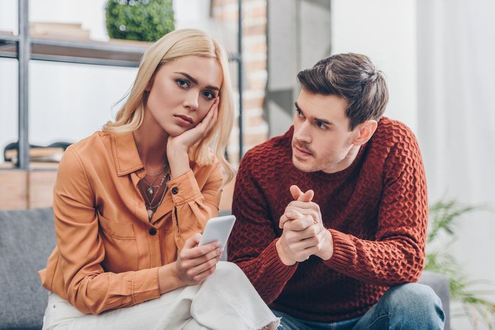 zodiak husband looking at unhappy young woman holding smartphone and looking at camera at home, relationship problem concept