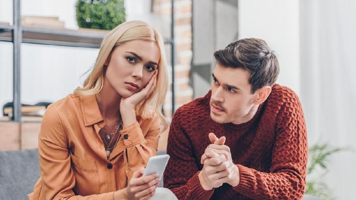 zodiak husband looking at unhappy young woman holding smartphone and looking at camera at home, relationship problem concept