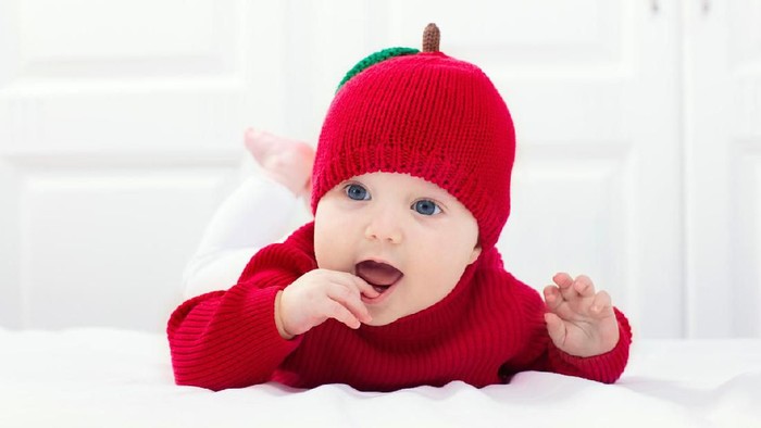 Baby in knitted apple hat. Kid on white bed. Child wearing red autumn sweater at home. Halloween or fall season costume.