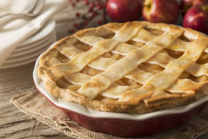 A top view of an apple pie with a slice removed on a white background.  The flaky, golden crust invites the viewer to reminisce.  The pie sits in a foil tin with one slice taken out leaving behind traces of the pie.  Pie filling just barely oozes out of the edges, leaving one to wonder who took the slice?