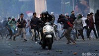 Seorang pengendara melintasi massa yang sedang melempari polisi dengan batu.