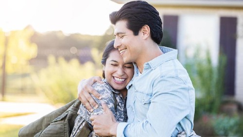 Young, Hispanic female in uniform hugs her husband tightly, after she has just arrived home from duty. She still holds her bag on her shoulder, while they stand outside their home.
