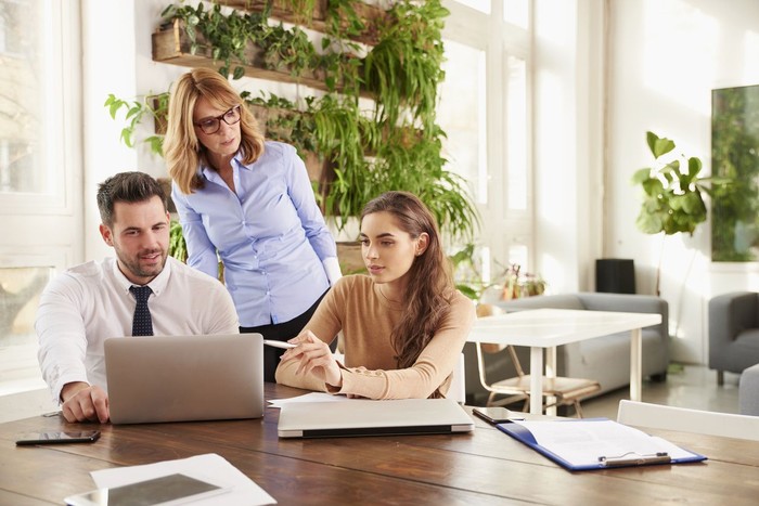 Group of business people working together on business project.  Young secretary woman and financial advisor businessman sitting in front of laptop while middle aged executive professional woman standing behind them and giving advice.