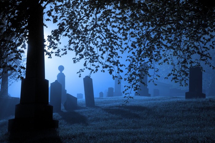 Gravestones and monuments are backlit by a bright light on a wet foggy evening.