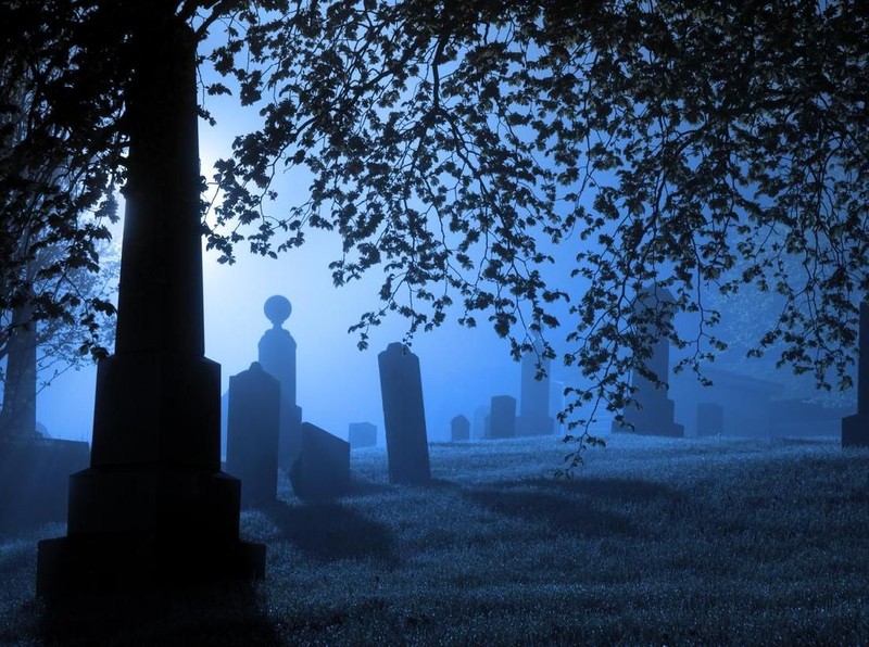 Gravestones and monuments are backlit by a bright light on a wet foggy evening.