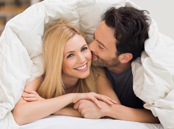 515600176 Handsome man kissing woman on cheek under a duvet in the bed. Happy young couple lying down comfortably in their bed at home. Young couple having fun lying on bed at home.