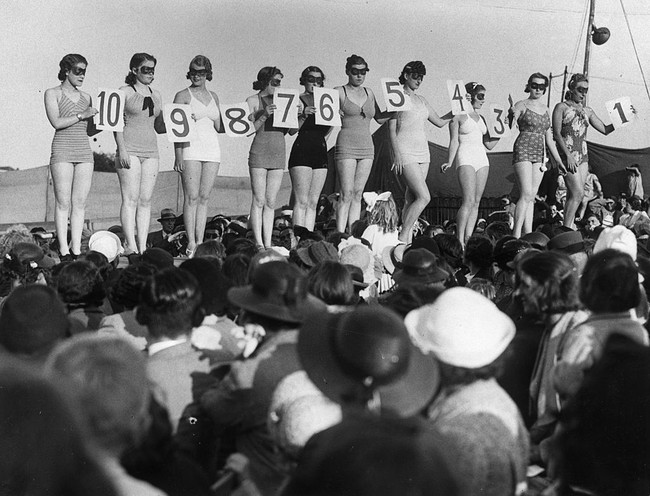 Pada zaman dahulu, peserta kontes kecantikan berparade di atas panggung dengan membawa papan nomer. Sedangkan di masa sekarang para finalis tampil dengan selempang yang menjadi identitas mereka. Biasanya pada selempang tertulis asal negara atau kota tempat tinggal mereka. Foto: Getty Images