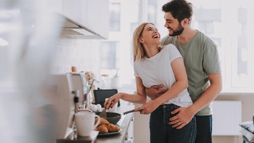 Cooked with love. Cute laughing blonde frying something in skillet. Her bearded husband is hugging her from behind