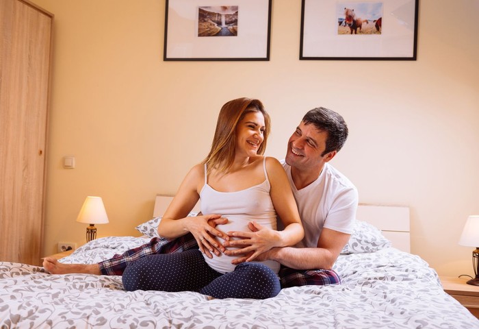 695893084 Adult couple - husband and pregnant wife sharing love in bed in the evening, hugging, kissing, holding baby bump and enjoying in their common happiness