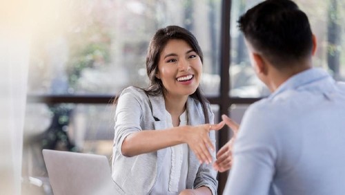 A smiling young businesswoman sits opposite an unrecognizable candidate during an interview.  She reaches across her desk to shake hands with him.