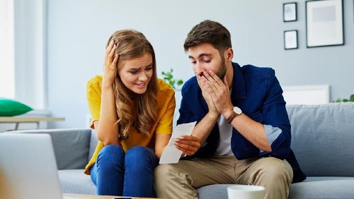Front view of a stressed young couple facing financial problems, sitting on couch and looking at bills