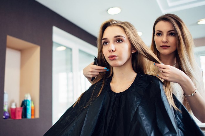 825115894 Hairstylist deciding with the young female customer what haircut to do in hairdressing parlor. Two pretty blondes discussing a new hairstyle