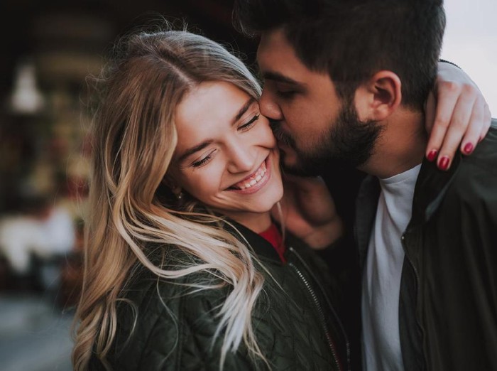 1086628458 Precious moments of love. Close up portrait of handsome bearded guy kissing his girlfriend in cheek while she hugging him. Lady closing eyes with pleasure and smiling