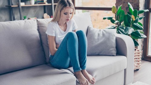 Upset woman has first symptoms of pms. She is sitting on a sofa and  touching her stomach. She is going to cry.