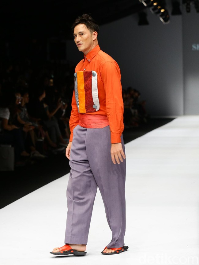 Disusul Mike Lewis, yang juga mengenakan rancangan Sebe11as, berupa kemeja oranye dengan aksen patchwork dipadu celana lebar abu-abu.  Foto: Rachman Haryanto/detikcom