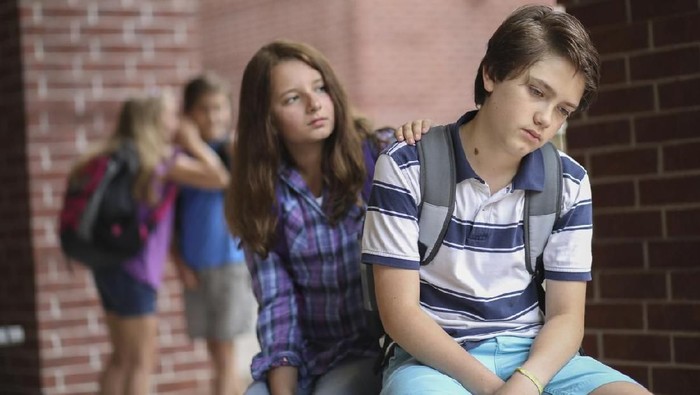 Friend comforts fellow student after he has been bullied outside the elementary school building,  Other students in background laugh at boy behind his back.