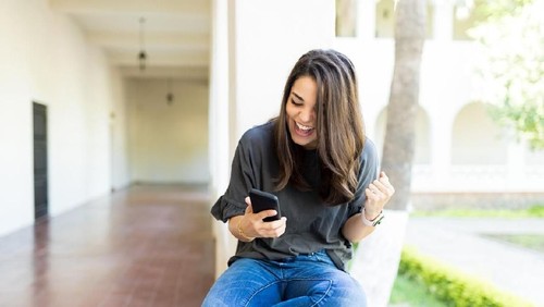 Excited female raising fist and cheering while watching sport on mobile phone in campus