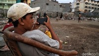 Sambil memagang gadget, seorang bocah menonton temannya bermain bola.